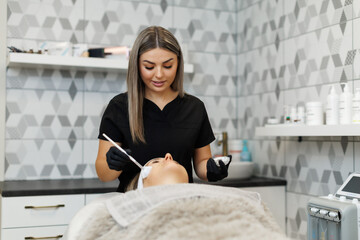 A smiling beautician in black uniform applies facial cream to a relaxed client using a brush. The spa setting includes skincare products and stylish tiled walls