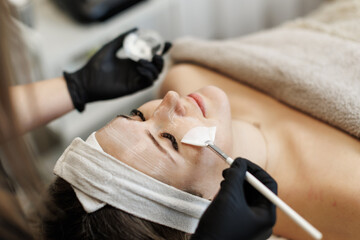 A woman lies peacefully as a beautician applies a clear cream mask with a brush to her face in a spa. Close-up view highlights skin texture and professional care