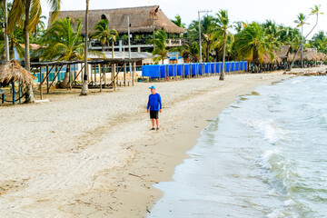 Caribbean man stands on the beach in Covenhas wearing a blue shirt and cap, surrounded by palm trees and coastal buildings with open copy space and calm tropical energy.