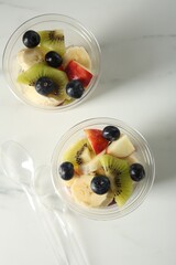 Tasty fruit salad in plastic cups and spoons on white marble table, flat lay