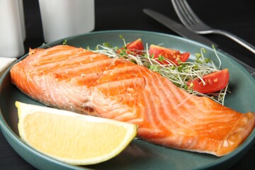 Piece of tasty grilled salmon, lemon, tomato slices and microgreens served on black table, closeup