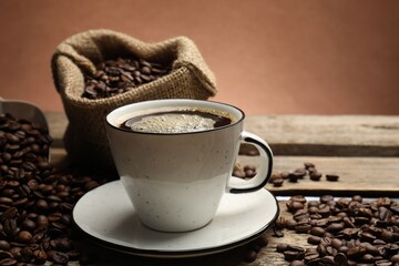 Aromatic coffee in cup and beans on wooden table against brown background, closeup. Space for text