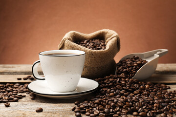Aromatic coffee in cup and beans on wooden table against brown background, closeup