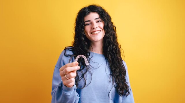 Happy young woman with long curly hair holding and pointing at a transparent removable aligner for dental correction on a yellow background