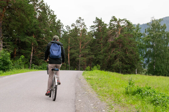 man with blue backpack riding bicycle on road surrounded by green trees and mountains in summer, view from behind