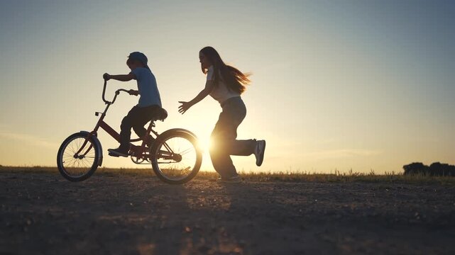 A boy rides a bike as a woman runs behind on the dirt path. The boy keeps pedaling the bike forward while the woman follows under the evening sky.