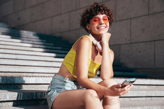 Young beautiful smiling hipster woman in trendy summer clothes. Carefree woman with curls hairstyle, in the street. Positive model holds mobile phone, looks at cellphone screen, uses smartphone apps