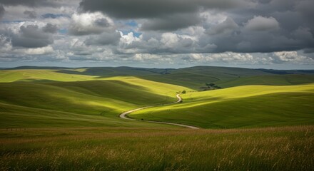 Obraz premium Winding country road carves through dramatic sunlit green rolling hills beneath a moody sky