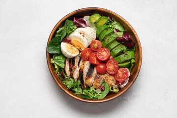 Healthy Cobb salad with grilled chicken breast, avocado, cherry tomatoes, lettuce and boiled egg in wooden bowl, top view on light background