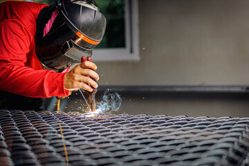 Welder wearing a red long-sleeved shirt and a protective mask is welding a steel grating,...