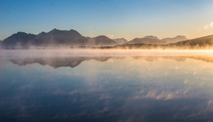 Misty lake reflects mountains at sunrise with golden light.