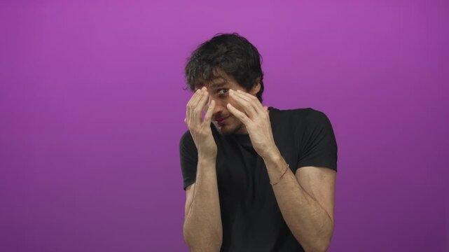 Young man raising hands defensively in studio with purple wall and tense expression and hunched posture; anxiety.