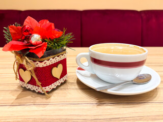A festive Christmas table setting with a cup of coffee beside a small holiday decoration with red flowers, ornaments, and wooden hearts.
