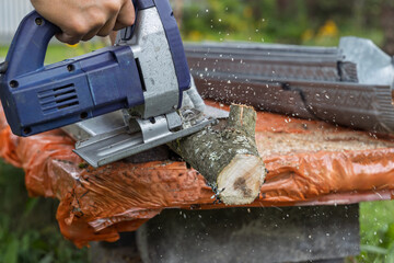 A person in work gloves uses a blue electric jigsaw to cut a tree branch. Wood shavings fly as the blade slices through the wood. Ideal for DIY, woodworking, and outdoor tool demonstration visuals