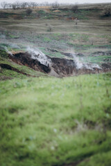 A rugged hillside emits white smoke from multiple cracks in the earth, creating a dramatic and surreal atmosphere.