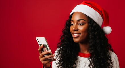 Smiling woman wearing a Santa hat and festive Christmas sweater, happily engaging with her smartphone on a vibrant red background, celebrating the holiday season