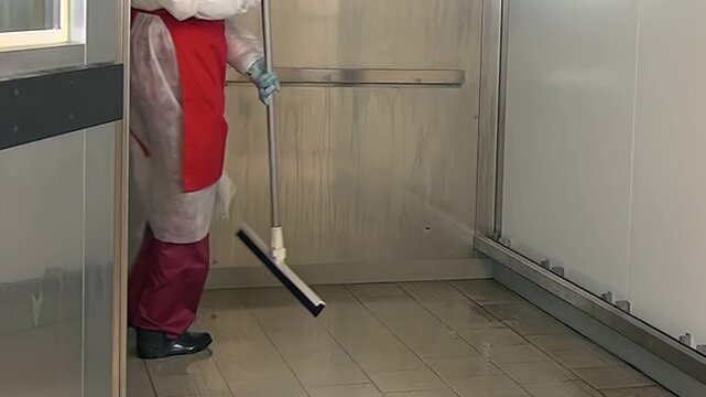 In what appears to be a sterile industrial setting, someone wearing protective gear and a red apron uses a floor squeegee to remove standing water from a tiled floor.