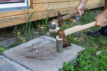 Person uses axe to chop small log placed on concrete slab outdoors. Scene includes wooden cabin wall, grass, and scattered firewood. Demonstrates tool use in backyard or rural setting
