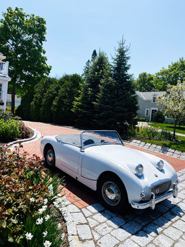 White vintage convertible Austin-Healey Sprite parked on brick driveway in a New England suburban yard