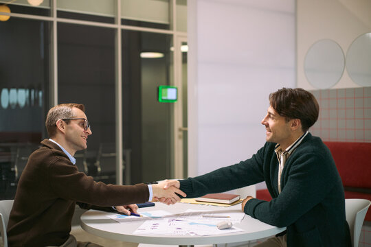 Two young adult Caucasian men sitting at round table shaking hands during business meeting, smiling and making eye contact, documents and digital tablet on tabletop