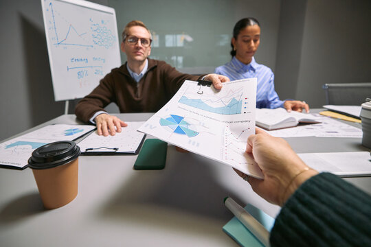Caucasian middle aged man and young adult Asian woman, sitting at desk analyzing financial charts while receiving business report from hand of unseen colleague during meeting