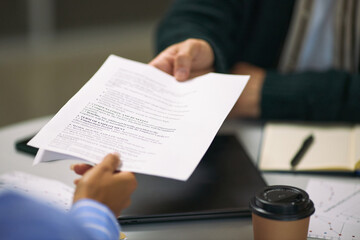 Caucasian middle aged man handing business document to Asian young adult man during meeting, both sitting at table with laptop, notebook and takeaway coffee cup visible in office setting