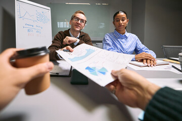 Caucasian middle aged man and Black young adult woman sitting at conference table reviewing business charts, while two colleagues handing documents and coffee during meeting