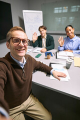 Portrait of young Caucasian man taking selfie with multiethnic young adult colleagues waving at camera during business meeting in modern office conference room