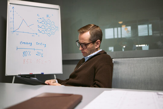 Caucasian middle aged man wearing glasses sitting at desk, analyzing data on digital tablet with business charts and diagrams on whiteboard in modern office meeting room