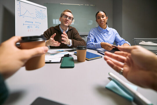 Caucasian middle aged man and young adult Asian woman discussing business strategy at meeting table with coffee cups, digital devices and documents, two people gesturing in foreground