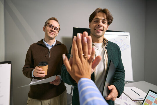 Caucasian young adult man and Caucasian middle aged man standing in office, smiling while greeting with high five, holding documents and coffee, business meeting in progress