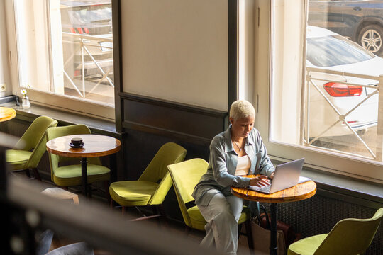 African American woman with white hair sits at desk with laptop, is surfing the Internet. female blogger, student, online education. lady works in coffee shop or coworking, top view.