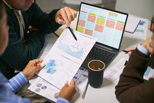 Three young adult men discussing financial charts and business analytics at desk, analyzing printed graphs and laptop screen with data, collaborating on project in office setting
