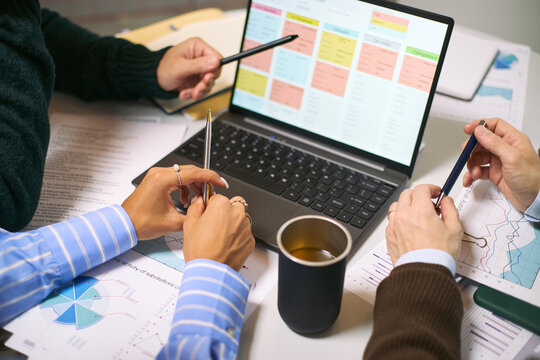 Three middle aged Caucasian and Asian women collaborating at desk, analyzing business charts and graphs, discussing data strategy with laptop and printed reports in office setting