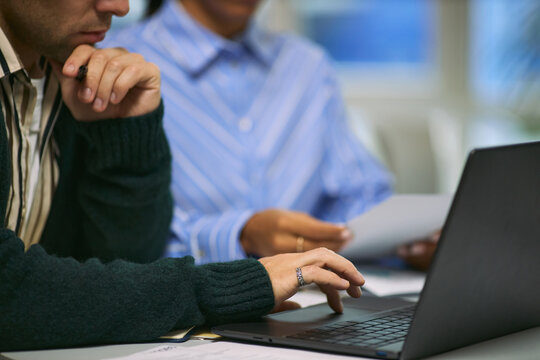 Caucasian young adult man working on laptop while sitting beside Black young adult woman reviewing documents during business meeting in modern office setting