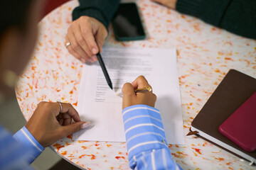 Caucasian woman and Black woman discussing business contract at round table, both reviewing printed document with pen and smartphone visible, focusing on agreement details