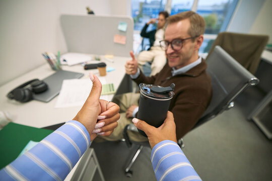 Caucasian middle aged man sitting at office desk giving thumbs up gesture while smiling, foreground showing hand of Black woman holding reusable coffee cup and giving thumbs up