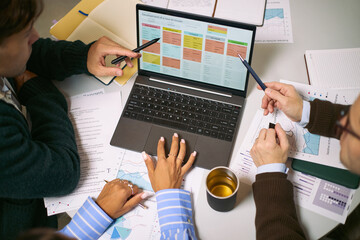 Diverse group of young adult and middle aged professionals collaborating around laptop, analyzing business charts and discussing project strategy in modern office setting