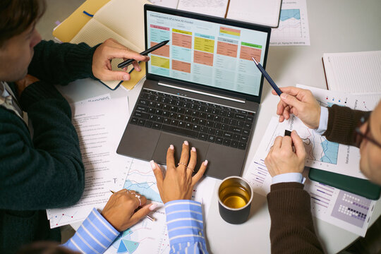 Diverse group of young adult and middle aged professionals collaborating around laptop, analyzing business charts and discussing project strategy in modern office setting - Powered by Adobe
