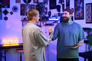 Caucasian young adult man with tattoos shaking hands with tattooed Caucasian middle aged man wearing beanie in tattoo studio, both smiling and making eye contact