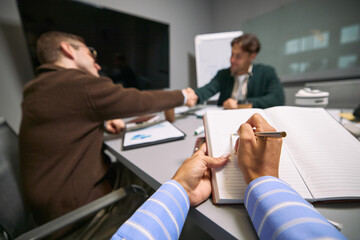 Young adult Caucasian man and young adult man shaking hands during business meeting while young adult Black woman taking notes in notebook, modern office setting, teamwork concept