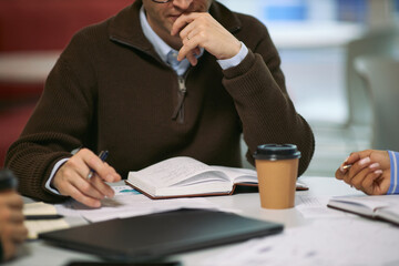 Middle aged Caucasian man sitting at desk, analyzing documents and writing notes in notebook during business meeting with disposable coffee cup and laptop on table