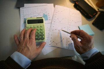 Middle aged Caucasian man analyzing financial charts with calculator and pen, reviewing business activity graphs and data sheets on desk, focusing on business performance metrics