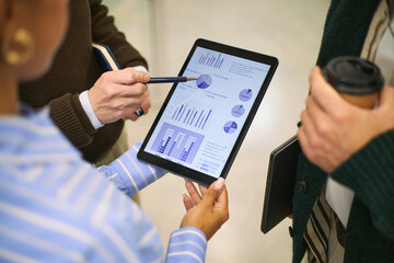 Caucasian man and Black woman analyzing financial charts on digital tablet during business meeting, holding stylus and coffee cup, discussing data visualization and performance metrics