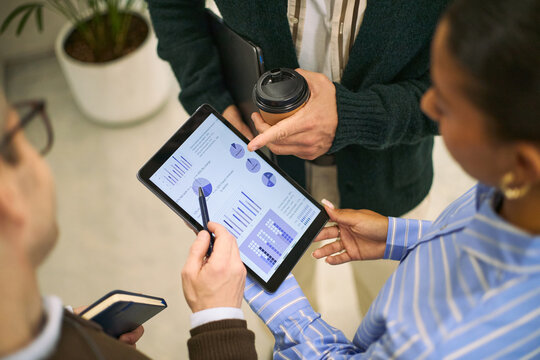 Young adult Caucasian man and young adult Black woman discussing financial charts on digital tablet during business meeting, both standing and collaborating with colleague holding notebook