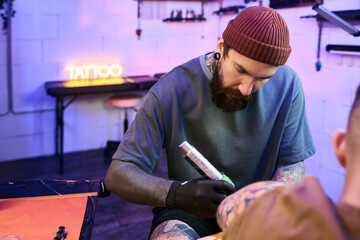 Caucasian young adult man with beard and tattoos working as tattoo artist, concentrating while creating tattoo on arm of another Caucasian young adult man in modern studio