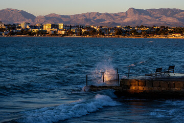 Fototapeta premium Sunlit waves crash against a stone pier at sunset, creating dramatic spray against the dark blue sea, with Side's coastal landscape and mountains glowing in golden evening light. Antalya, Turkey.