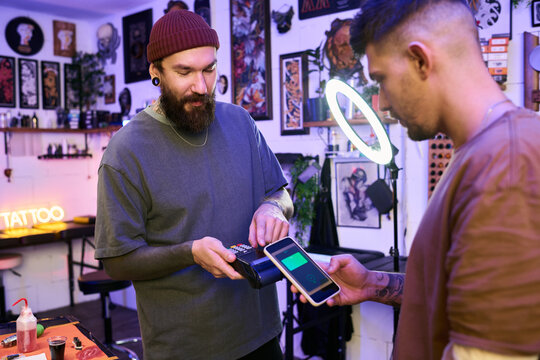 Caucasian young adult man with beard processing mobile payment on card reader while another Caucasian young adult man holding smartphone in tattoo studio