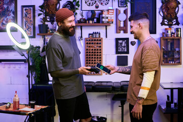 Two young adult Caucasian men standing in tattoo studio exchanging smartphones, both smiling and making eye contact, tattoo equipment and artwork visible in background