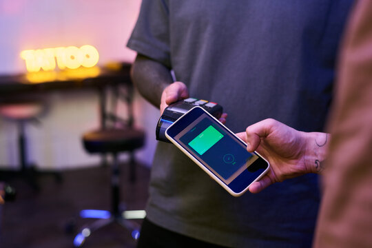 Caucasian young adult man holding smartphone making contactless payment using mobile wallet app while another young adult man holding card reader in tattoo studio setting
